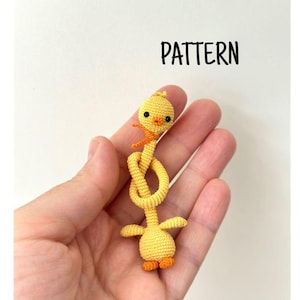 May include: A handmade yellow crochet duck with orange feet and a small orange scarf. The duck's body is elongated and knotted, with the word "PATTERN" in black text above the duck.
