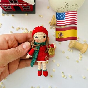 Peut inclure: Poupée au crochet faite à la main, vêtue d'un manteau, d'un chapeau et de bottes rouges, avec une écharpe verte. La poupée tient un petit panier. Elle mesure environ 10 cm. En arrière-plan, une petite maison rouge, des drapeaux et des éléments décoratifs.