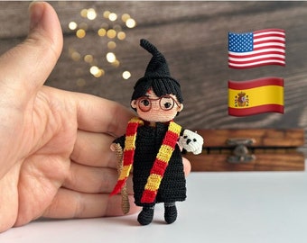 May include: A crocheted Harry Potter doll wearing a black robe, red and yellow scarf, and round glasses. The doll is holding a wand and a small white owl. The doll is sitting on a wooden surface with an American and Spanish flag in the background.
