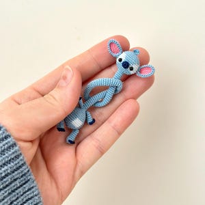 May include: A light blue crochet figure with pink-lined ears, a dark blue nose, and white accents. The figure is in a knotted pose. The figure is a handmade item.
