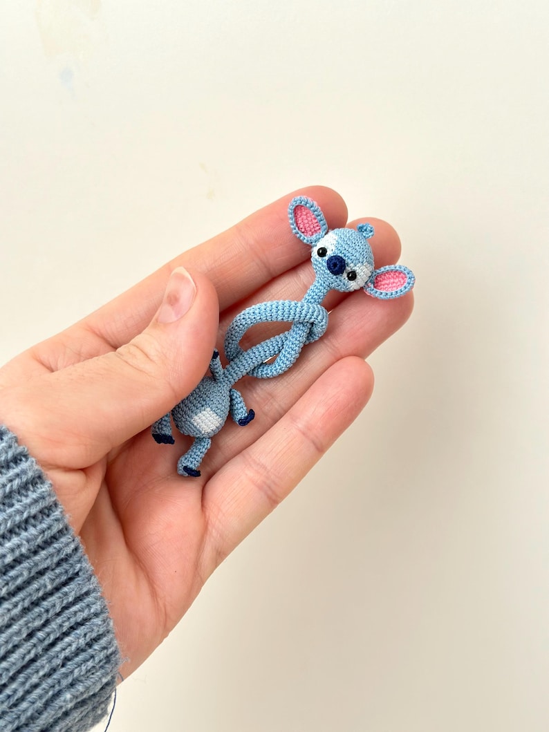 May include: A light blue crochet figure of a cartoon character with large ears, a dark blue nose, and pink inner ears. The figure is held in a hand, showcasing the detailed crochet work. The figure is approximately 3 inches in length.