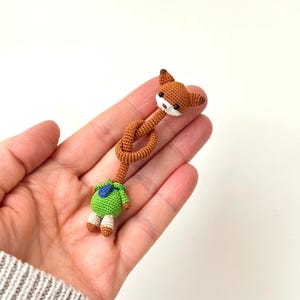 May include: A handmade crochet fox doll with a knotted body. The fox has brown fur, white muzzle, and black eyes. It wears a green outfit with a blue accent. The doll is held in a person's hand.