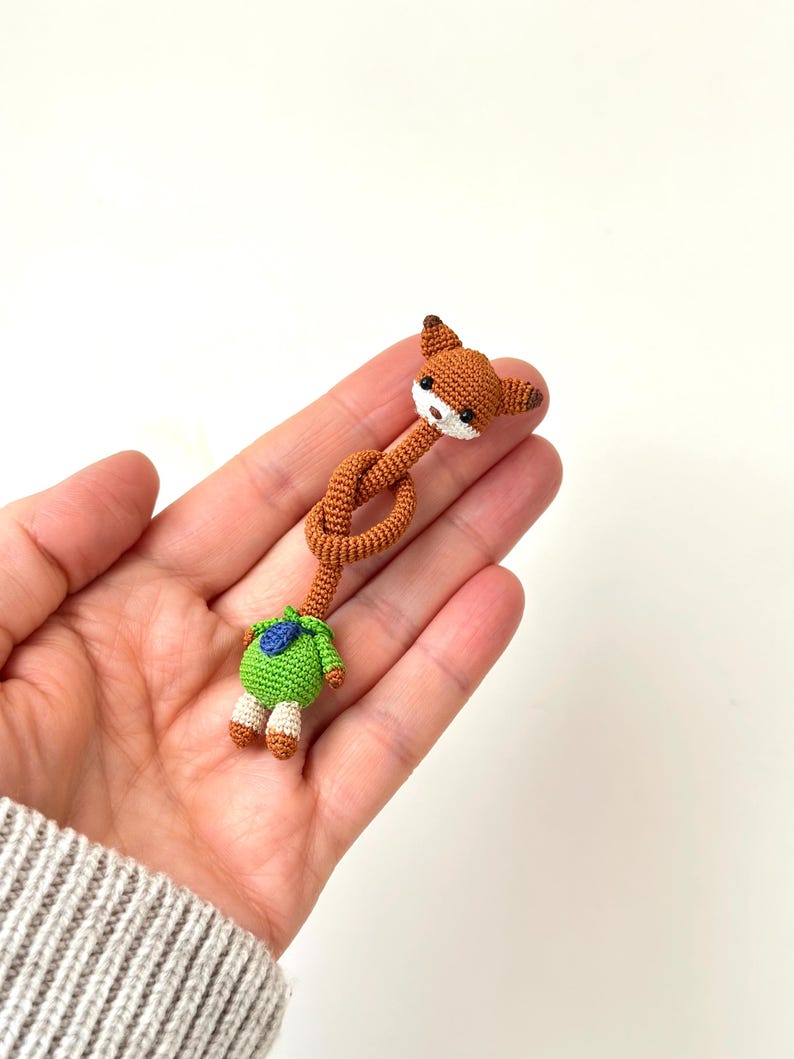 May include: A handmade crochet fox doll with a knotted body. The fox has brown fur, white muzzle, and black eyes. It wears a green outfit with a blue accent. The doll is held in a person's hand.