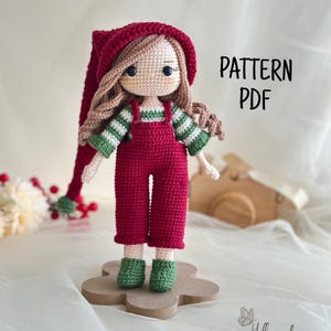 Peut inclure: Poupée au crochet faite à la main, vêtue d'une tenue rouge, de chaussures vertes et d'un chapeau assorti. La poupée a de longs cheveux bruns et se tient sur une base en bois. Le texte "PATTERN PDF" est visible.