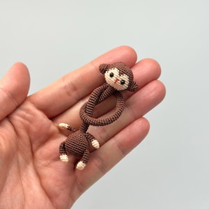 May include: A small, handmade crochet monkey in shades of brown and cream. The monkey has a detailed face with black eyes and a red mouth. Its arms and legs are attached to a looped body, creating a playful design. A charming, handcrafted item.