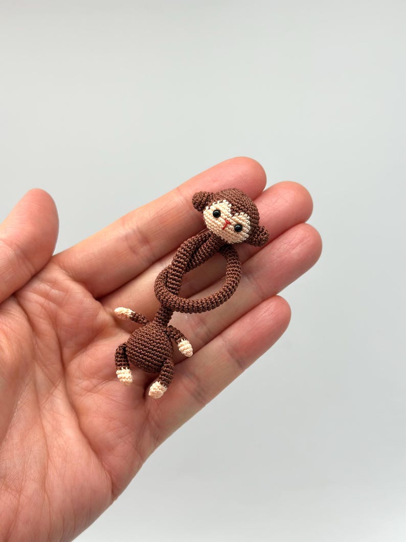 May include: A small, handmade crochet monkey in shades of brown and cream. The monkey has a detailed face with black eyes and a red mouth. Its arms and legs are attached to a looped body, creating a playful design. A charming, handcrafted item.