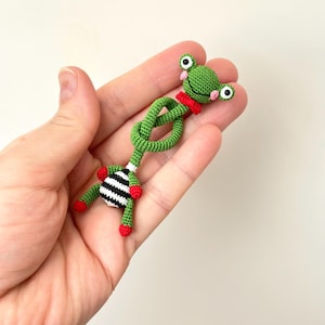 May include: A handmade green crochet frog with a red bow tie and black and white striped legs. The frog has large, round eyes and red feet. The frog is a whimsical, handcrafted item.