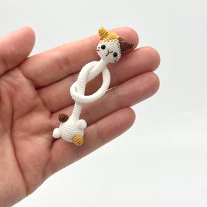 May include: A handmade crochet cat figurine in white, brown, yellow, and black. The cat's head is detailed with small ears and facial features. The body is a knotted design, and the cat is held in a hand.