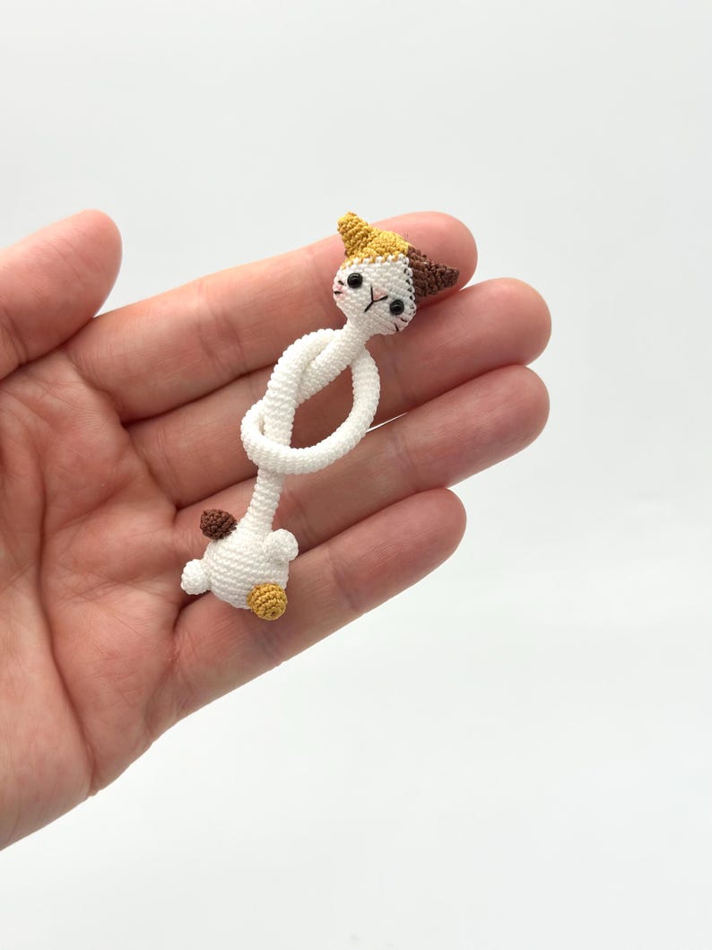 May include: A handmade crochet cat figurine in white, brown, yellow, and black. The cat's head is detailed with small ears and facial features. The body is a knotted design, and the cat is held in a hand.