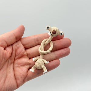 May include: A beige crocheted alien-like figure with large black eyes, a brown nose, and a smiling mouth. The figure's neck is knotted, and it has small arms and legs with white tips. The toy is held in a hand against a white background.