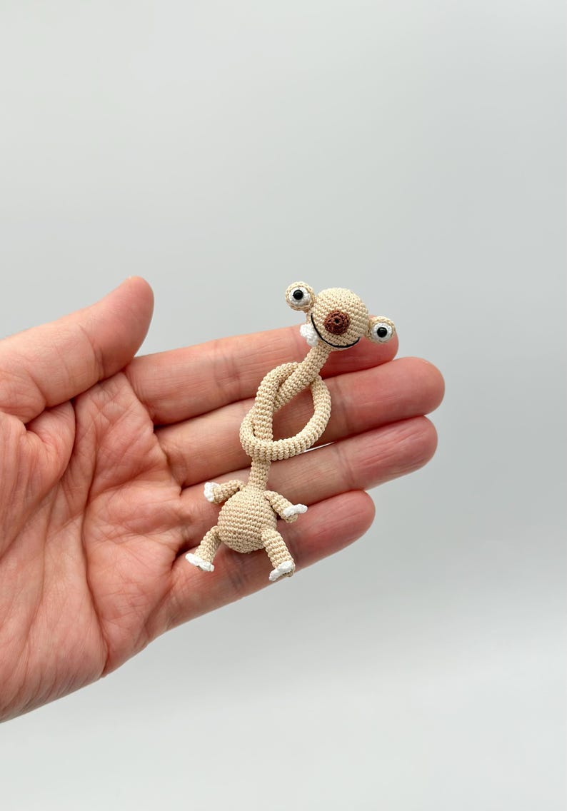 May include: A beige crocheted alien-like figure with large black eyes, a brown nose, and a smiling mouth. The figure's neck is knotted, and it has small arms and legs with white tips. The toy is held in a hand against a white background.