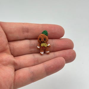 Op de afbeelding: Een miniatuur gehaakte peperkoeken man figuur. De figuur is bruin met een groene hoed, witte en rode accenten op de voeten en handen, en een geel en rood ontwerp op de torso. De figuur wordt in een hand gehouden.
