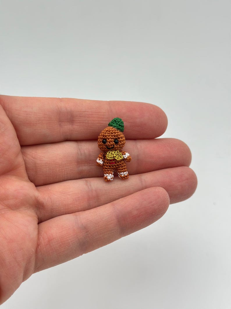 Op de afbeelding: Een miniatuur gehaakte peperkoeken man figuur. De figuur is bruin met een groene hoed, witte en rode accenten op de voeten en handen, en een geel en rood ontwerp op de torso. De figuur wordt in een hand gehouden.