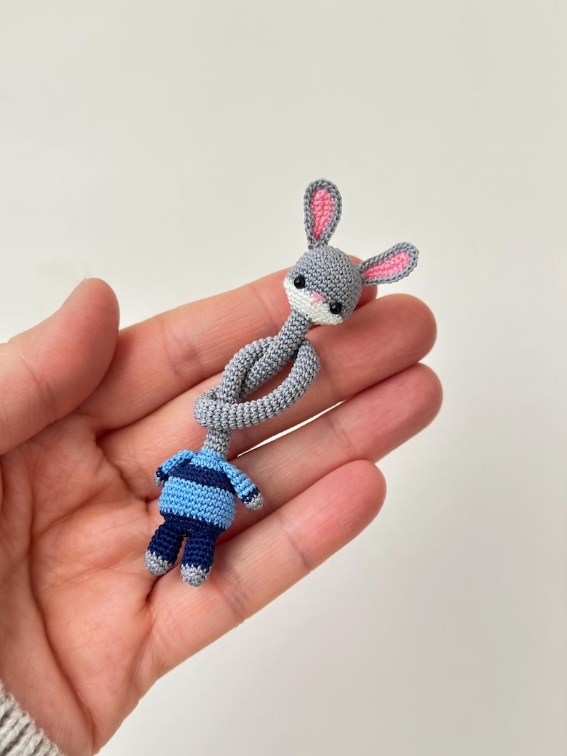 May include: A handmade crochet bunny doll with a gray head and pink-lined ears. The bunny's body is blue and navy, with a striped shirt and matching pants. The doll is held in a hand against a white background.
