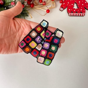 May include: A small, colorful crocheted vest with a granny square pattern. The vest is made with black yarn and features squares in various colors, including red, green, blue, yellow, and pink.