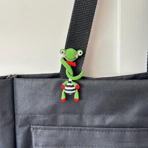 May include: A handmade crochet frog charm in green, red, and black and white stripes, attached to a black bag strap. The frog has large eyes and a red bow tie. The bag is black with a zipper and a small charm.