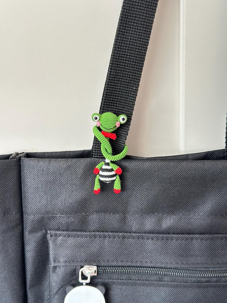 May include: A handmade crochet frog charm in green, red, and black and white stripes, attached to a black bag strap. The frog has large eyes and a red bow tie. The bag is black with a zipper and a small charm.