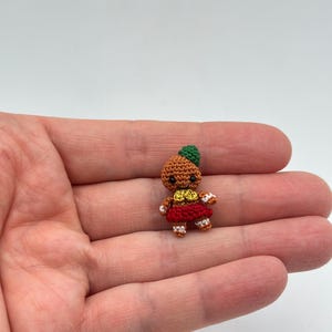 Op de afbeelding: Een kleine, handgemaakte gehaakte pop met een bruin lichaam, groene hoed, rode rok en witte accenten. De pop wordt in een hand gehouden, wat de kleine afmetingen en ingewikkelde details benadrukt. Het gezicht van de pop heeft een lachende uitdrukking.