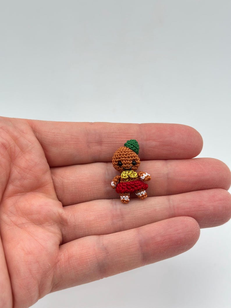 Op de afbeelding: Een kleine, handgemaakte gehaakte pop met een bruin lichaam, groene hoed, rode rok en witte accenten. De pop wordt in een hand gehouden, wat de kleine afmetingen en ingewikkelde details benadrukt. Het gezicht van de pop heeft een lachende uitdrukking.