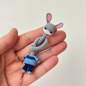 May include: A handmade crochet bunny doll. The bunny has a gray head with pink-lined ears and a blue and navy body. The doll is wearing a striped shirt and matching trousers. The doll is held in a hand.