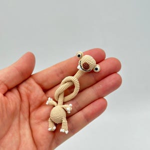 May include: A crocheted beige alien figure with large black eyes and a brown mouth. The alien has a long, knotted neck and small arms and legs with white accents. The figure is held in a hand against a white background.