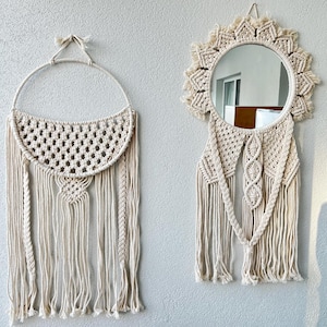 Puede incluir: Dos colgantes de pared de macramé en color blanco roto natural. Uno es un diseño de semicírculo con un patrón tejido y flecos. El otro es un espejo redondo con un marco de macramé en forma de sol y flecos colgantes.