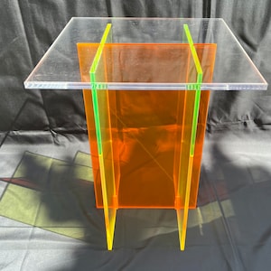 May include: A clear acrylic table with a bright orange base and neon green accents. The table has a square top and four legs.
