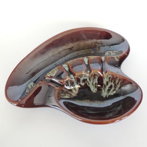 May include: A brown and black ceramic ashtray with a unique, organic shape. The ashtray has a glossy finish with areas of light green and brown detailing. The design includes multiple indentations for resting cigarettes.