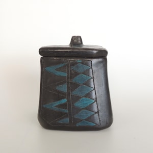 May include: A dark gray ceramic jar with a lid. The jar features a geometric design of teal blue diamonds. The jar is square with a slightly tapered shape and a small handle on the lid.
