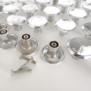 May include: A collection of vintage cabinet knobs with a shiny chrome finish. The knobs have a round, flat top and a cylindrical base. Several screws are also visible, suggesting they are ready for installation. The knobs are arranged on a white surface.