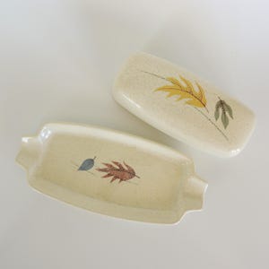 May include: Two vintage ceramic ashtrays with a speckled cream background. One is rectangular with rounded edges and a small handle on each side. The other is a rectangular lid. Both have leaf designs in yellow, brown, and blue.