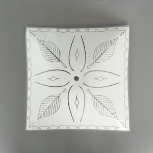 May include: Square white ceiling light fixture with a black leaf and diamond design. The fixture has a scalloped edge and a central hole. The design is symmetrical, with four leaves and diamond shapes radiating from the center.