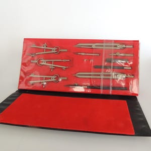 May include: A set of silver drafting tools, including compasses and dividers, neatly arranged in a red case. The case is open, revealing the tools against a red velvet-like interior. The exterior is black.