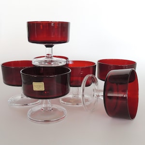 May include: A collection of six ruby red glass dessert bowls. The bowls have a wide, shallow design with a clear stem and base. One bowl is lying on its side. The bowls are arranged on a white surface.
