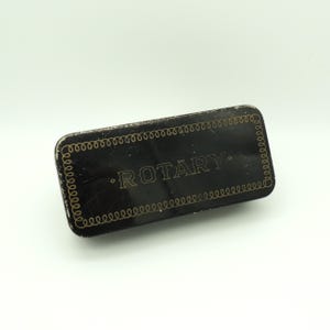 May include: A rectangular black metal box with the word "ROTARY" in gold lettering. The box has a decorative gold swirl border. The box is slightly scratched and worn, suggesting it is vintage or antique.