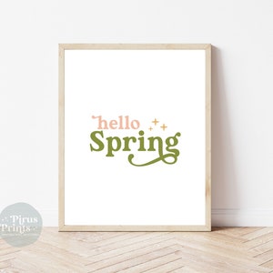 Op de afbeelding: Een ingelijst print met de tekst "hello spring" in roze en groene letters met gouden sterren. De print is op een witte achtergrond en is ingelijst in een licht houten frame.