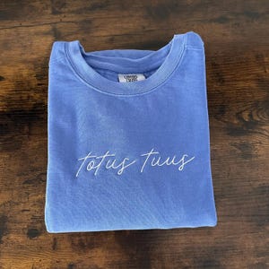 May include: A periwinkle blue crew neck sweatshirt with the embroidered text "Totus Tuus" in a cursive font. The sweatshirt is folded neatly on a dark wood surface. The brand tag "Comfort Colors" is visible at the neckline.