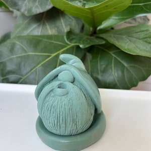 May include: A sage green garden gnome figurine with a long beard and pointed hat. The gnome is sitting on a circular base. The background features large green leaves.