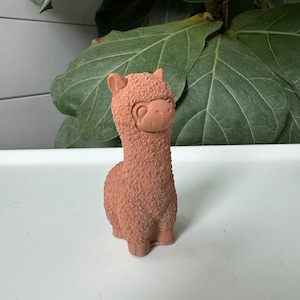 Puede incluir: Una figurita de llama de cerámica marrón con una superficie texturizada, que se asemeja al pelaje. La llama tiene una cara linda con ojos negros y una boca pequeña.