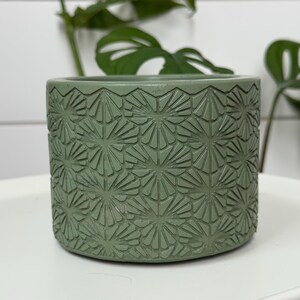 Può includere: Un vaso cilindrico verde salvia con un motivo geometrico dettagliato e ripetuto. Il piccolo vaso rotondo è realizzato in un materiale strutturato ed è adatto a piccole piante. Il vaso è appoggiato su una superficie bianca.