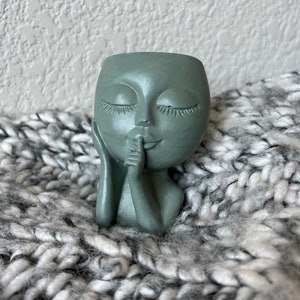 Puede incluir: Una maceta en forma de rostro verde salvia con los ojos cerrados y un dedo tocando los labios en un gesto de 'shush'. La maceta decorativa pequeña está hecha de un material liso y descansa sobre una manta de punto gris y blanco. Ideal para plantas pequeñas.