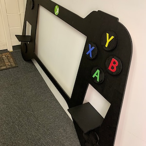 May include: Black wooden cutout of a video game controller with shelves on the sides. The controller has a green X button, a blue X button, a red A button, and a yellow Y button.