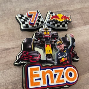 May include: A red, white, and blue checkered racing flag with the number 7 on it. A red bull logo is on the other flag. A Formula 1 race car with a driver in a red and blue racing suit is in the center of the image. The driver is holding up his right hand in victory. The name Enzo is written in orange letters with glitter on a black background.
