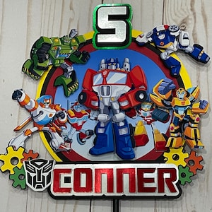 May include: A Transformers themed cake topper with a red, blue, and yellow robot in the center. The robot is surrounded by other smaller robots in green, blue, and yellow. The number 5 is in the top center of the topper. The name "CONNER" is written in red letters at the bottom of the topper.