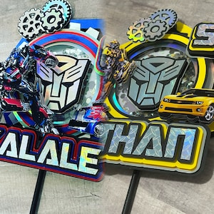 May include: Two Transformers themed cake toppers with the names "Alale" and "Ethan" in iridescent letters. The toppers feature a blue and red color scheme with a robot and car, and a yellow and black color scheme with a robot and car.
