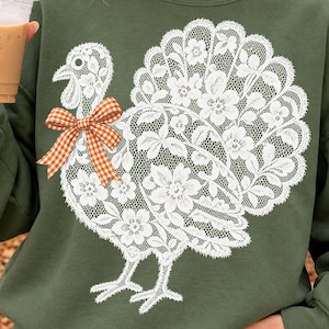 May include: An olive green crewneck sweatshirt featuring a large white graphic of a turkey composed of intricate floral lace patterns. The turkey design includes a prominent orange and white checkered bow tie accent on its neck area.