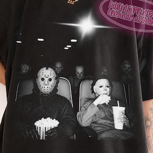 Peut inclure: Un t-shirt noir &agrave; manches courtes avec une impression graphique de personnages de films d'horreur assis dans un cin&eacute;ma. L'image montre des personnages masqu&eacute;s, dont Jason Voorhees et Michael Myers, tenant du pop-corn et un gobelet.