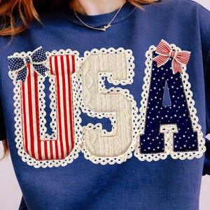 May include: Navy blue sweatshirt with the word "USA" appliqued on the front. The letters are decorated with red and white stripes, a cable knit pattern, and a navy blue star pattern. Each letter is outlined with white lace and topped with a bow.