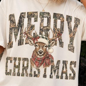 Puede incluir: Una camiseta de manga corta color hueso con el texto MERRY CHRISTMAS en estampado de camuflaje. Centrado entre las palabras hay una ilustración de un ciervo con un gorro de punto verde y blanco y una bufanda de cuadros rojos y verdes.