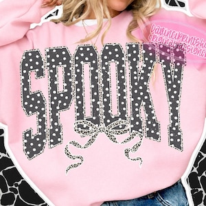 May include: A light pink long-sleeve sweatshirt featuring the word SPOOKY in large, dark gray block letters with white polka dots. Below the text is a decorative bow graphic with a black and white leopard print pattern. The text DavinciPrintShop is visible.